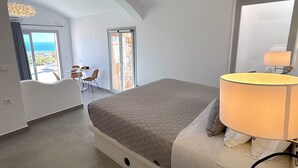 Een slaapkamer met een bed, een lamp, uitzicht door een deur, en een zithoek met een tafel en stoelen.