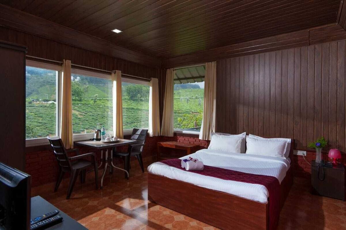A hotel room with a bed, a desk, and a chair. There are windows with a view of greenery outside.