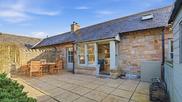 A stone cottage with a tiled patio, a dining area with wooden furniture, and a sign reading 'The Beach.' - 5 Courtyard Cottages - North East Escapes (Embleton)