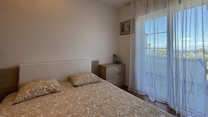Quarto com cama, mesa de cabeceira, cômoda, janela com cortinas e luminária fixada na parede.