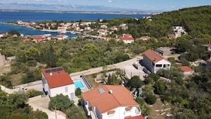 Vista aérea de uma área residencial costeira com casas, piscina e vista para o mar e montanhas ao fundo.