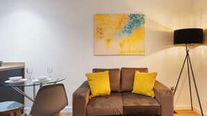 Ein modernes Wohnzimmer mit einem Glastisch, zwei Sofas mit gelben Kissen, einer Stehlampe und einem Gemälde an der Wand.