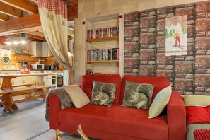 Una sala acogedora con un sofá rojo, estantes de madera con libros, y una cocina al fondo.