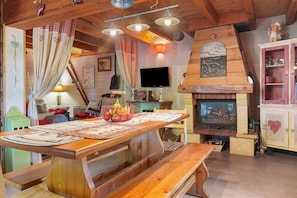 Un interior rústico con una mesa de comedor de madera, bancos, una chimenea, un televisor y un mueble con puertas de vidrio.
