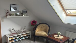 A room with a sloped ceiling, a bookshelf, a chair, a small table, and a window with blinds. - Apartment 'Ferienwohnung Hühnergott' with Wi-Fi (Sassnitz)