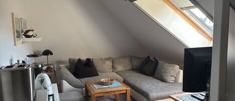 A loft-style living room with a sloped ceiling, a sofa, a coffee table, a television, and a kitchenette area.