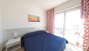 Un dormitorio con cama, mesita de noche, ventana con cortinas y un cuadro de un atardecer.