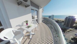 A balcony with a white table and chairs, a potted plant, and a view of the ocean and buildings. - Quiet beachside flat with sea view (Lignano Sabbiadoro)