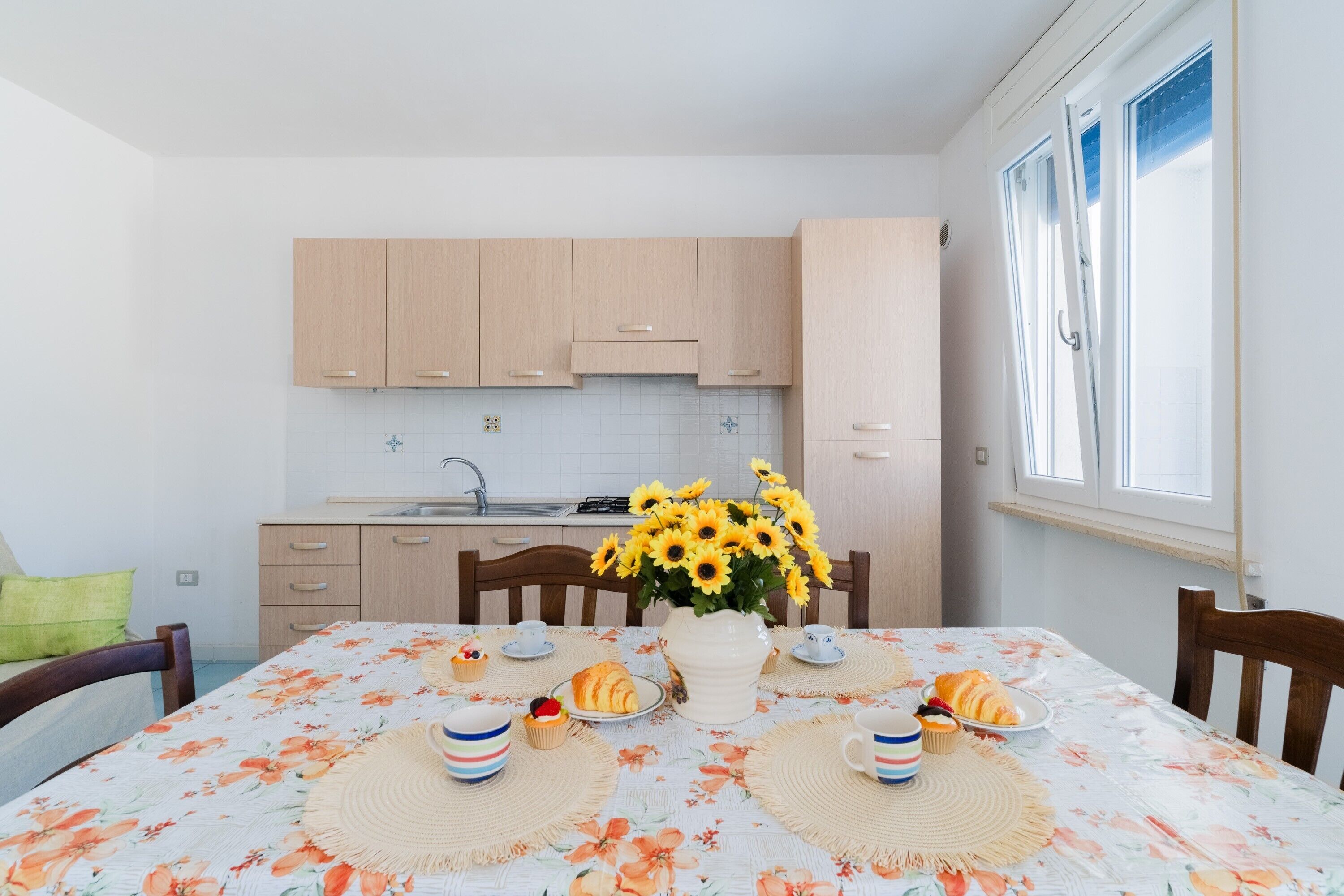 Une table à manger recouverte d’une nappe à motifs floraux, un vase rempli de tournesols et une table dressée pour le petit-déjeuner, avec des croissants et des muffins.