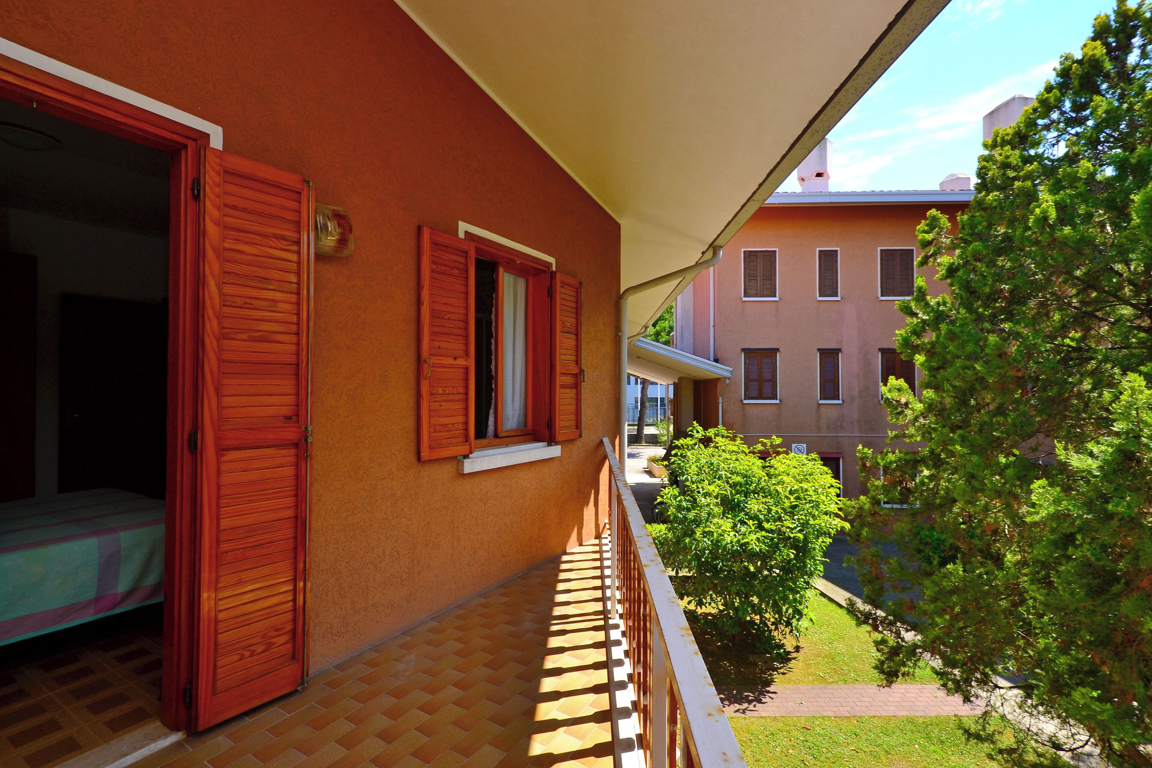 A balcony with a wooden door, a window with shutters, and a view of a building and greenery.