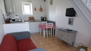 Una cocina compacta con microondas, tostadora y hervidor, una zona de comedor con mantel a cuadros rojo y blanco, un sofá gris y un televisor montado en la pared.