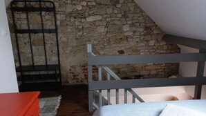 Una habitación con pared de piedra, escalera de madera, cama con colcha a rayas y cómoda roja.