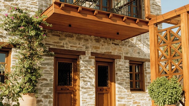 Ein zweistöckiges Steinhaus mit einem hölzernen Balkon, großen Kübelpflanzen und einer Holzpergola.