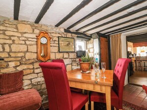 A dining area with a stone wall, wooden ceiling beams, a wooden table, red upholstered chairs, a wall-mounted clock, and a potted plant. - The Annexe at Cherry Cottage (Gloucester)