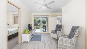 A compact room with a bed, a TV, a small cabinet, wicker chairs, a ceiling fan, and a sliding glass door leading to an outdoor area. - Welcome to your perfect family-friendly holiday retreat (Corlette)