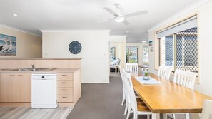 A modern kitchen with a dishwasher, a dining area with a wooden table and white chairs, a ceiling fan, and a clock on the wall. - Welcome to your perfect family-friendly holiday retreat (Corlette)