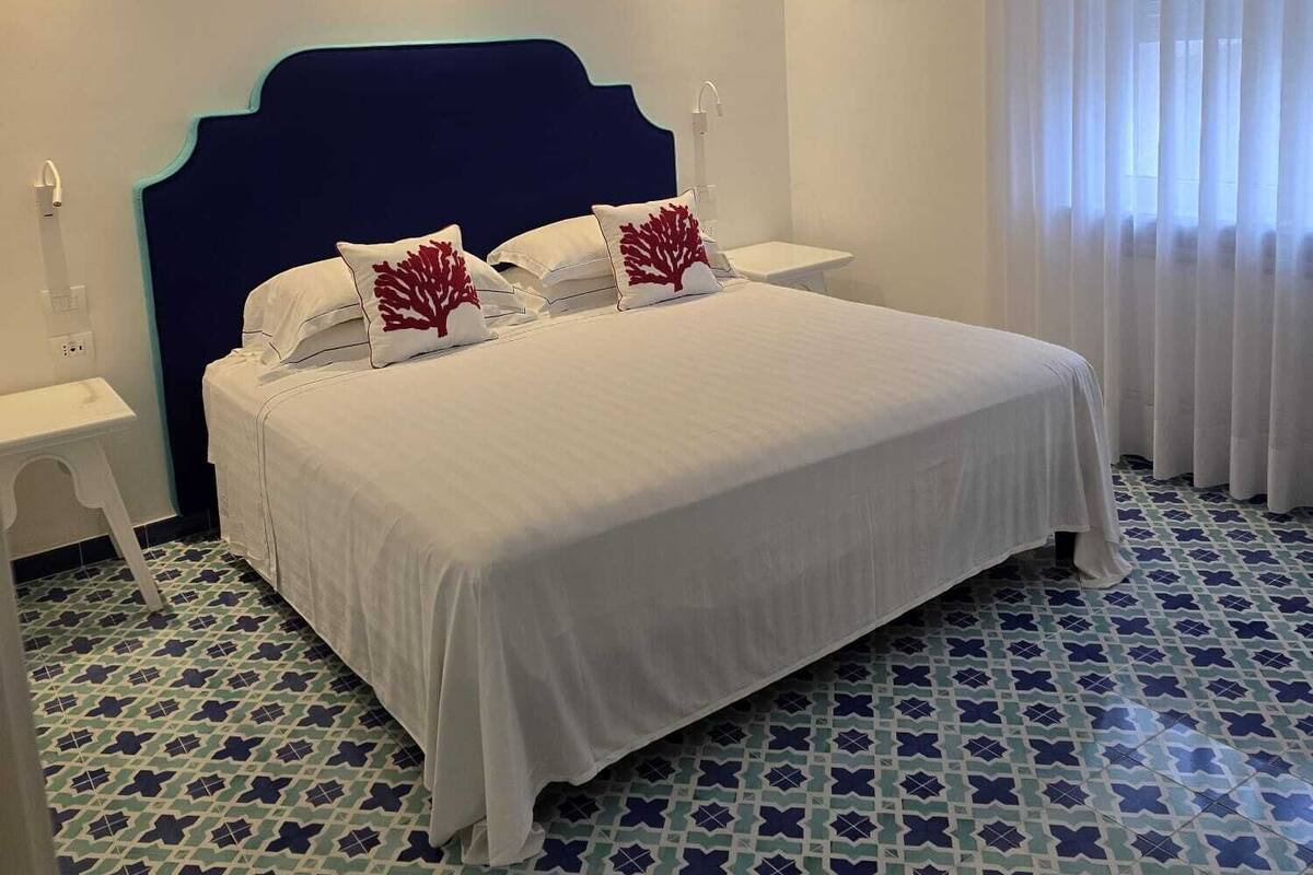 A bed with white bedding and red decorative pillows, a dark blue headboard, white bedside tables, and a patterned floor.