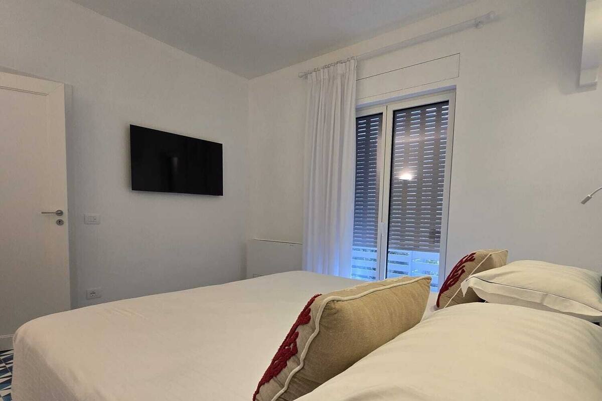 A hotel room with a bed, a flat-screen TV, a window with blinds, and a patterned floor.