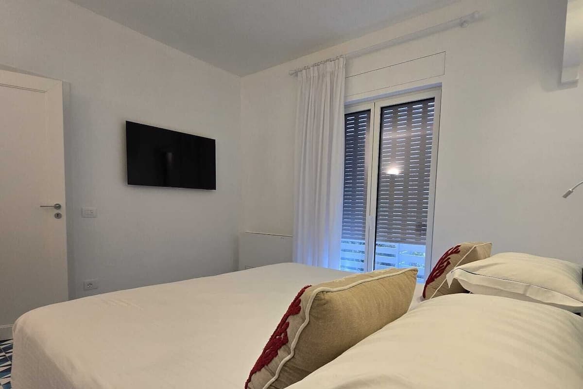 A hotel room with a bed, a flat-screen TV, a window with blinds, and a patterned floor.