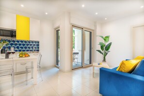 Ein modernes Wohnzimmer mit einem blauen Sofa, eine Küche mit einer gelben Akzentwand und eine Glastür, die zu einem Außenbereich führt.