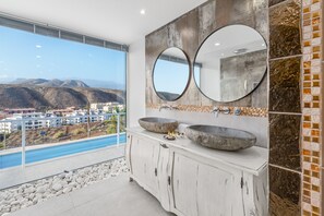 Ein Badezimmer mit einem großen Fenster, zwei runden Spiegeln, einem weißen Waschtisch mit zwei Steinbecken und Blick auf einen Pool und Gebäude.