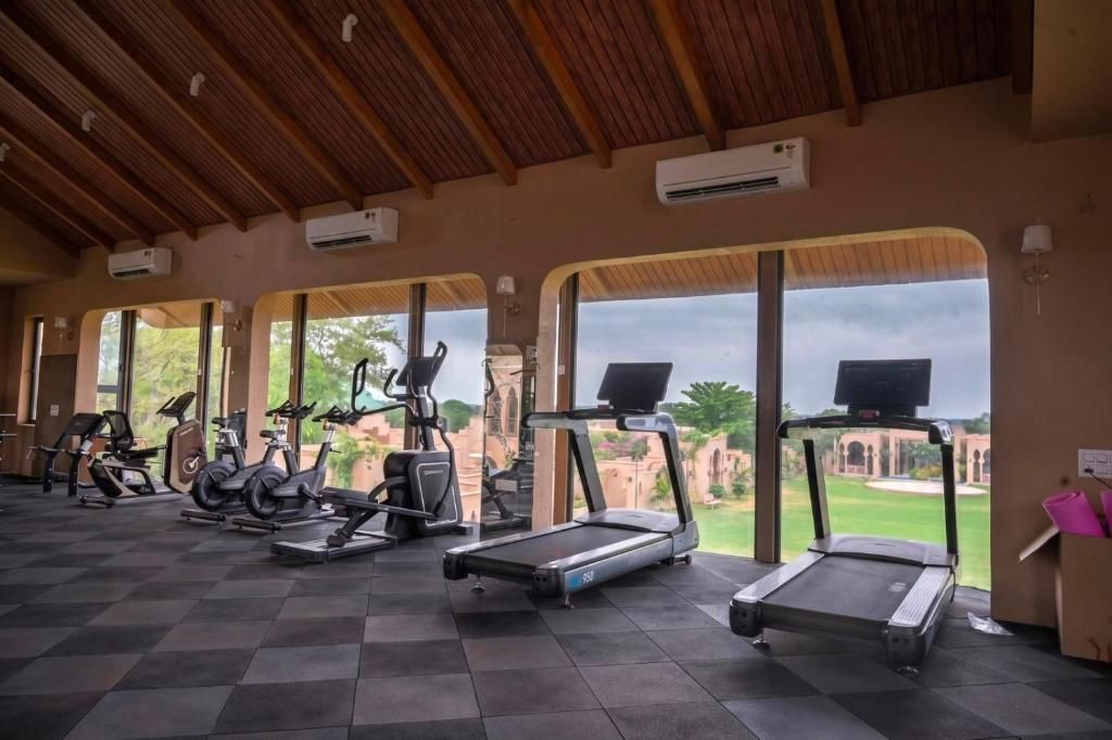 A gym with treadmills, stationary bikes, and elliptical machines, with large windows offering a view of greenery and architecture.