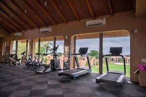 A gym with treadmills, stationary bikes, and elliptical machines, with large windows offering a view of greenery and architecture. - Nirva Club and Resort by Siara (Modasa)