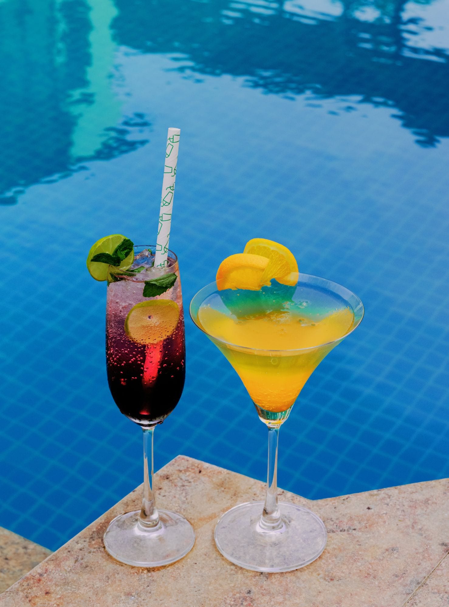 Two cocktails by a poolside with clear blue water in the background.