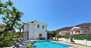 Une piscine avec des chaises longues, un bâtiment blanc doté d’un balcon, et un paysage montagneux en arrière-plan.