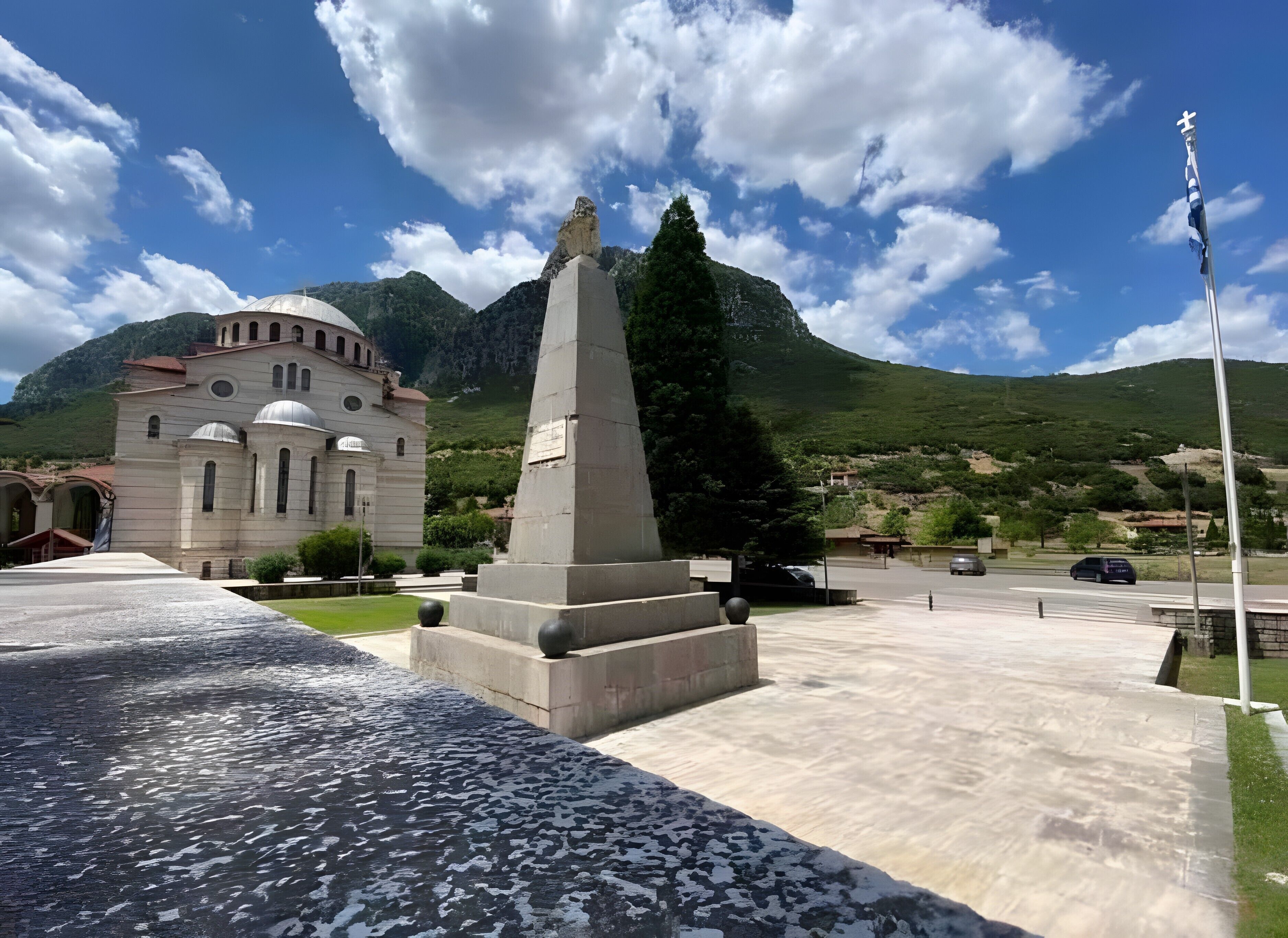 A monument with a cross, a church with domes, and mountains in the background.