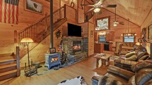 A log cabin interior with a stone fireplace, a wooden staircase, a dining area, and a living room with leather furniture. - A River Runs Through It by Chatuge Home Concierge | Private Riverfront · Rustic Luxury Cabin · Fireplace · Deck Views · Kayak & Fish Access (Hayesville)