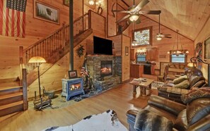 A log cabin interior with a stone fireplace, a wooden staircase, a dining area, and a living room with leather furniture. - A River Runs Through It by Chatuge Home Concierge | Private Riverfront · Rustic Luxury Cabin · Fireplace · Deck Views · Kayak & Fish Access (Hayesville)
