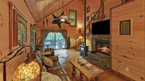 A cozy cabin interior with a stone fireplace, leather sofa, and a television. - A River Runs Through It by Chatuge Home Concierge | Private Riverfront · Rustic Luxury Cabin · Fireplace · Deck Views · Kayak & Fish Access (Hayesville)