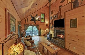 A cozy cabin interior with a stone fireplace, leather sofa, and a television. - A River Runs Through It by Chatuge Home Concierge | Private Riverfront · Rustic Luxury Cabin · Fireplace · Deck Views · Kayak & Fish Access (Hayesville)