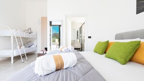 Quarto de hotel moderno com cama beliche, uma cama grande com almofadas verdes e laranja, cabeceira branca e vista através de uma porta aberta.