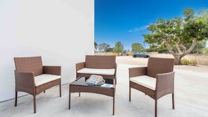 Um pátio com móveis de vime e uma mesa de vidro, céu azul claro e uma árvore ao fundo.