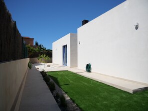 Ein modernes Gebäude mit weißer Fassade, einer blauen Tür und einem kleinen Gartenbereich mit Kunstgrashalm und Pflanzen.