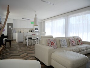 Een moderne woonkamer met een bank, een eettafel met stoelen, een keuken en grote ramen.