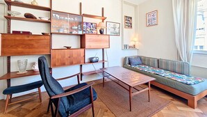 A living room with a sofa, a coffee table, a chair, shelves with decorative items, and a window with curtains. - New! N&J -LE CATINAT - Spacious - Balcony (Nice)