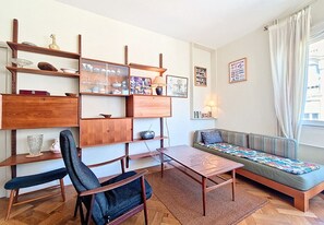 A living room with a sofa, a coffee table, a chair, shelves with decorative items, and a window with curtains. - New! N&J -LE CATINAT - Spacious - Balcony (Nice)