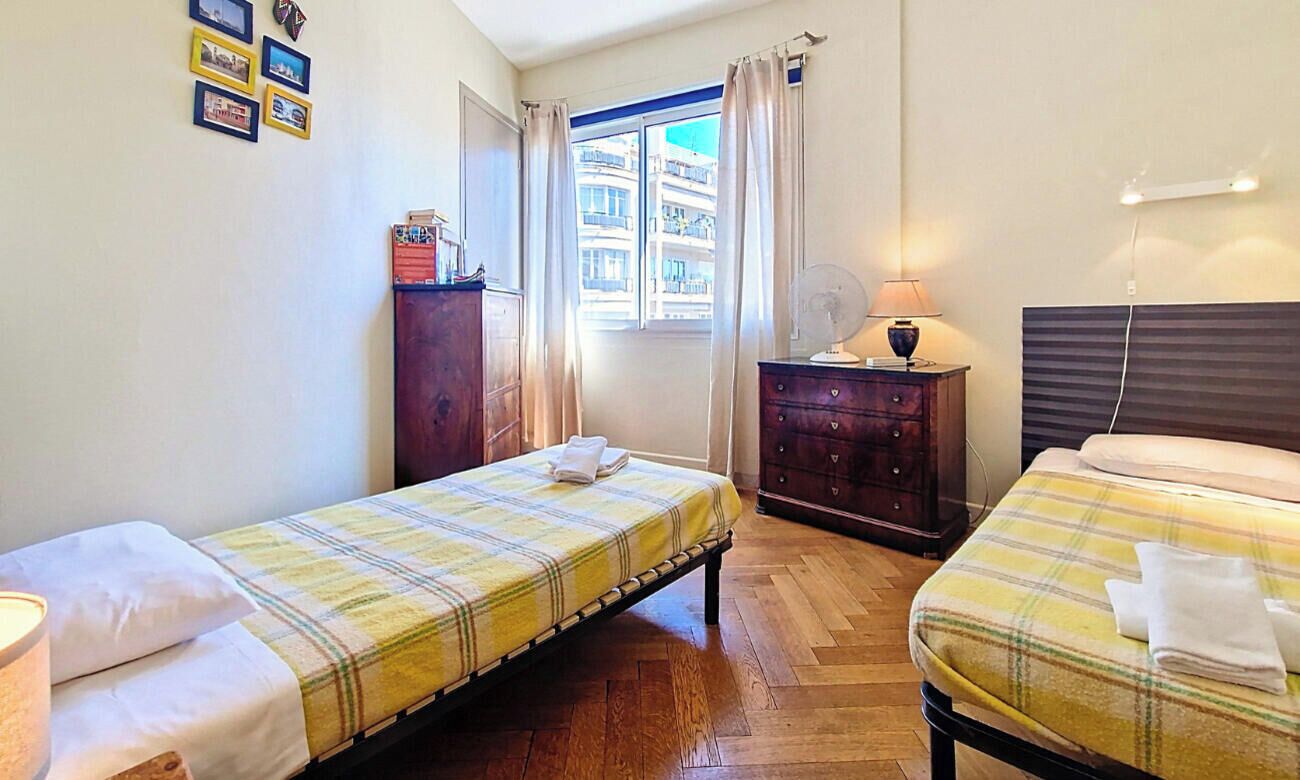 A room with two beds, a window with curtains, a dresser, and a wall-mounted fan.