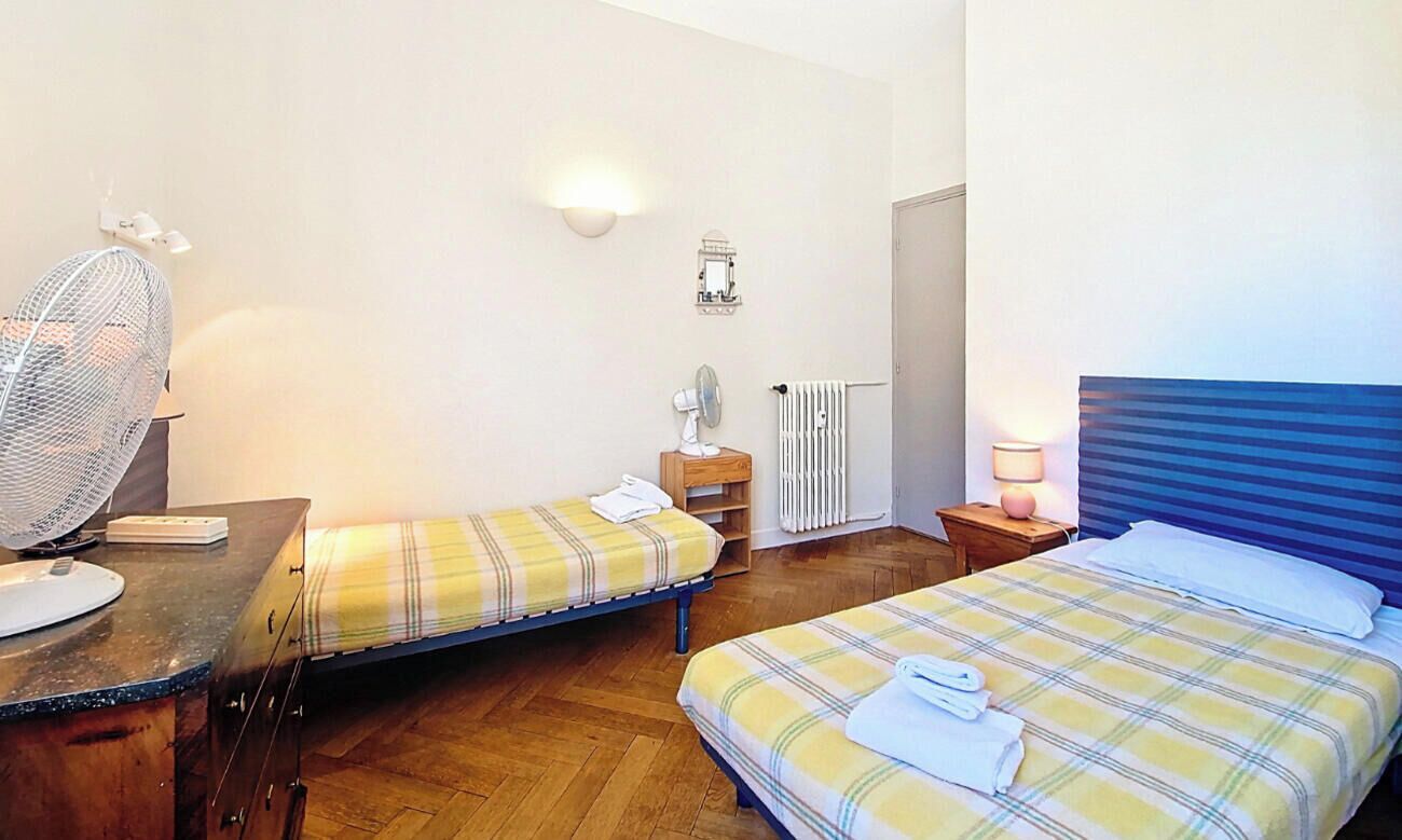 A bedroom with two beds, a desk, a chair, a fan, a radiator, and a wall-mounted light fixture.