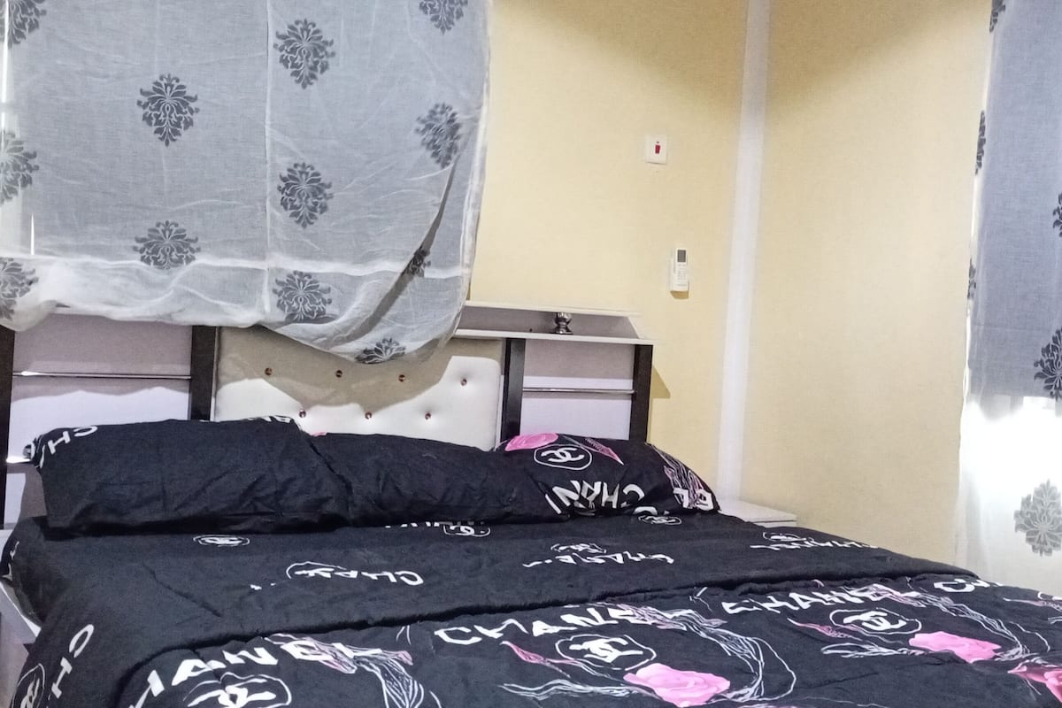Una cama con un edredón negro y rosa inspirado en Chanel, un cabecero blanco y una mesita de noche con una lámpara.