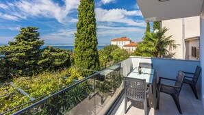 A balcony with a glass railing, a table, and chairs, overlooking a garden and a building with a red roof. - Modern vacation apartment with fantastic views and swimming pool (Crikvenica)