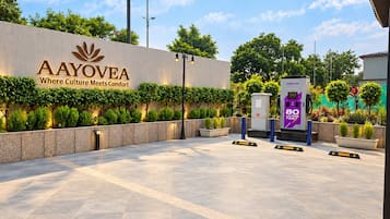 A modern entrance with a sign reading 'AAYOVEA Where Culture Meets Comfort,' a solar-powered electric vehicle charging station, and a well-maintained landscaped area.