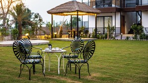A patio with two chairs and a table, a thatched umbrella, and a modern building in the background. - Aayovea Resort Ayodhya Near Ram Mandir & Saryu River (Ayodhya)