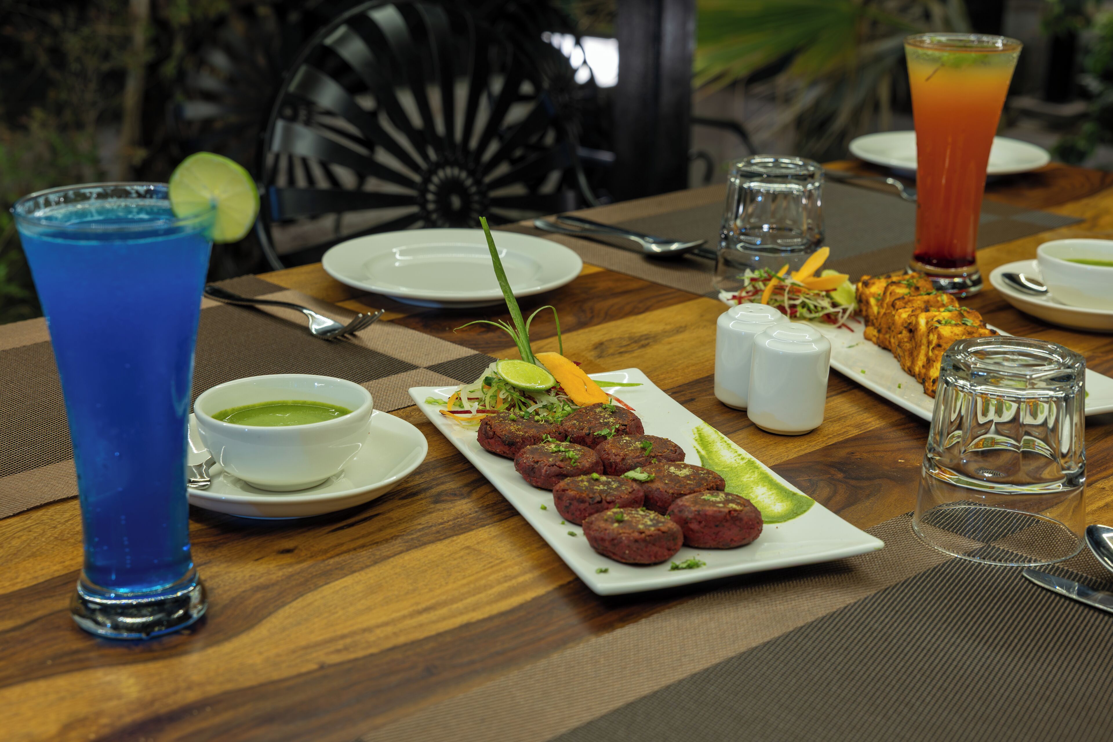 A dining table with a plate of food, a glass of blue drink, a bowl of green soup, and a tall glass of red drink.