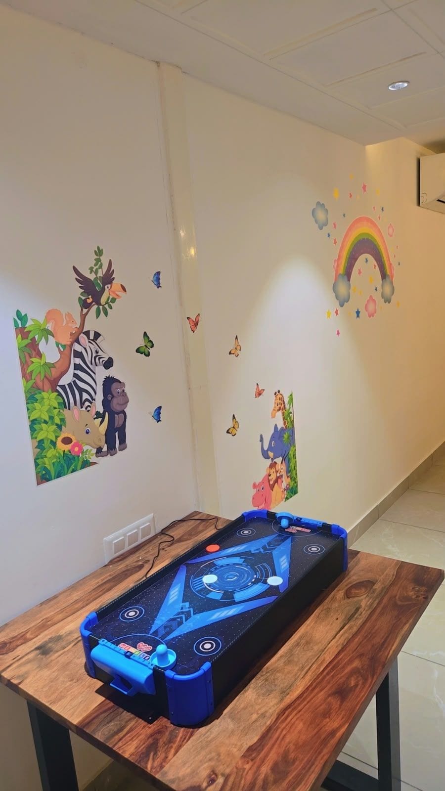 A room with a wooden table, a blue gaming table, and colorful wall decorations including animals and a rainbow.