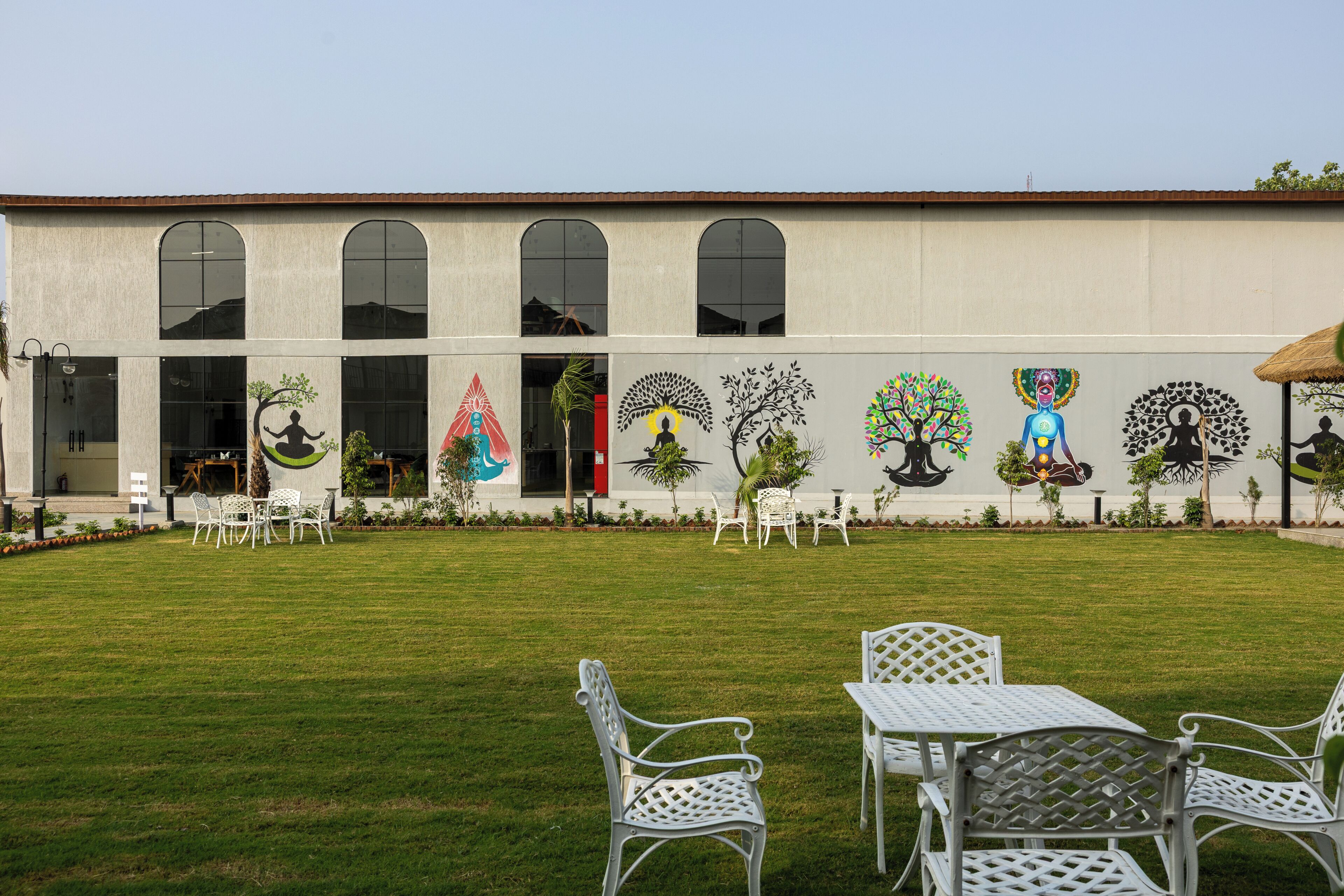 A building with large arched windows, outdoor seating, and a mural with tree and human figures.