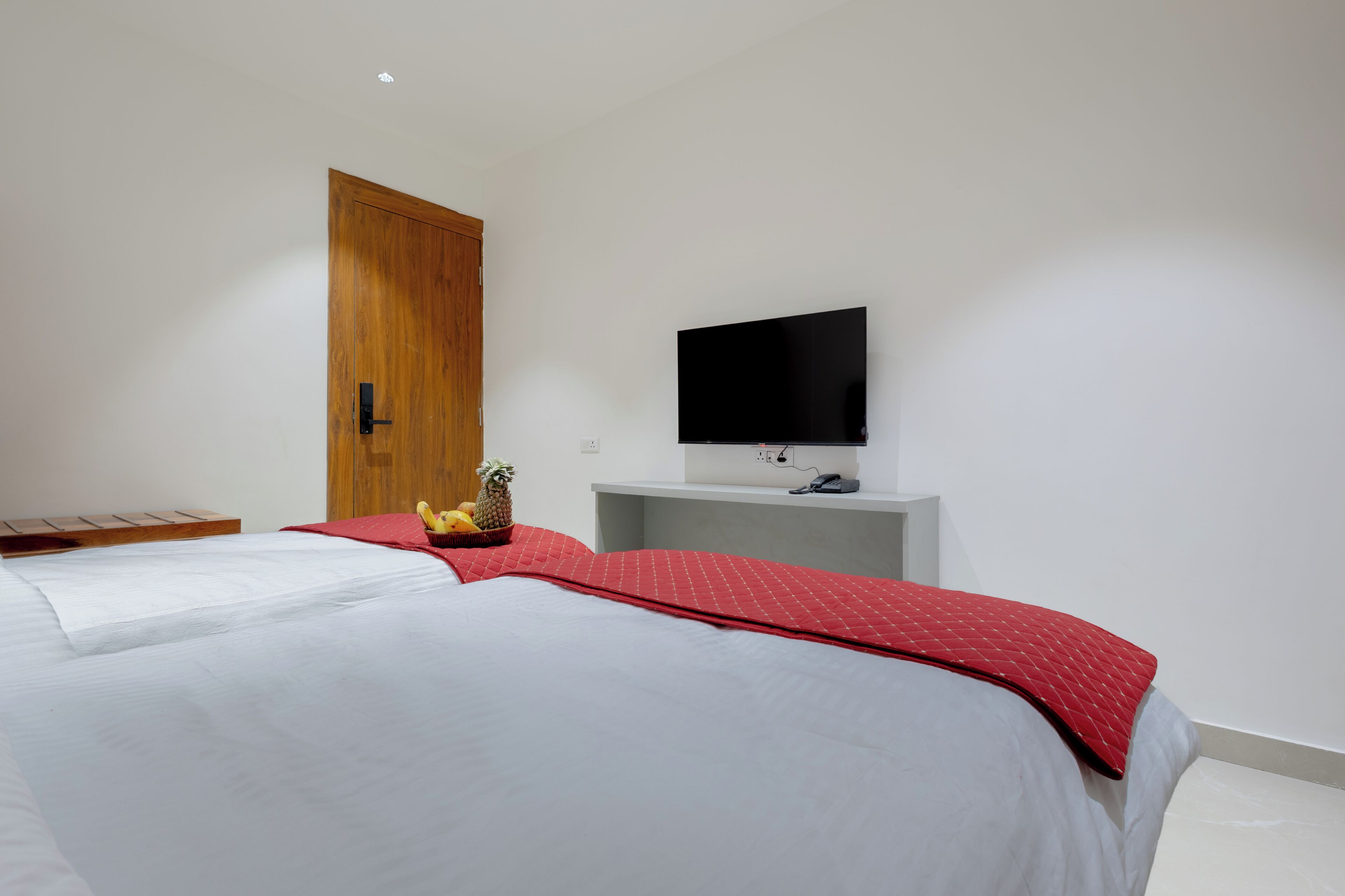 A bedroom with a bed, a television on a stand, a wooden wardrobe, and a small table with fruits.